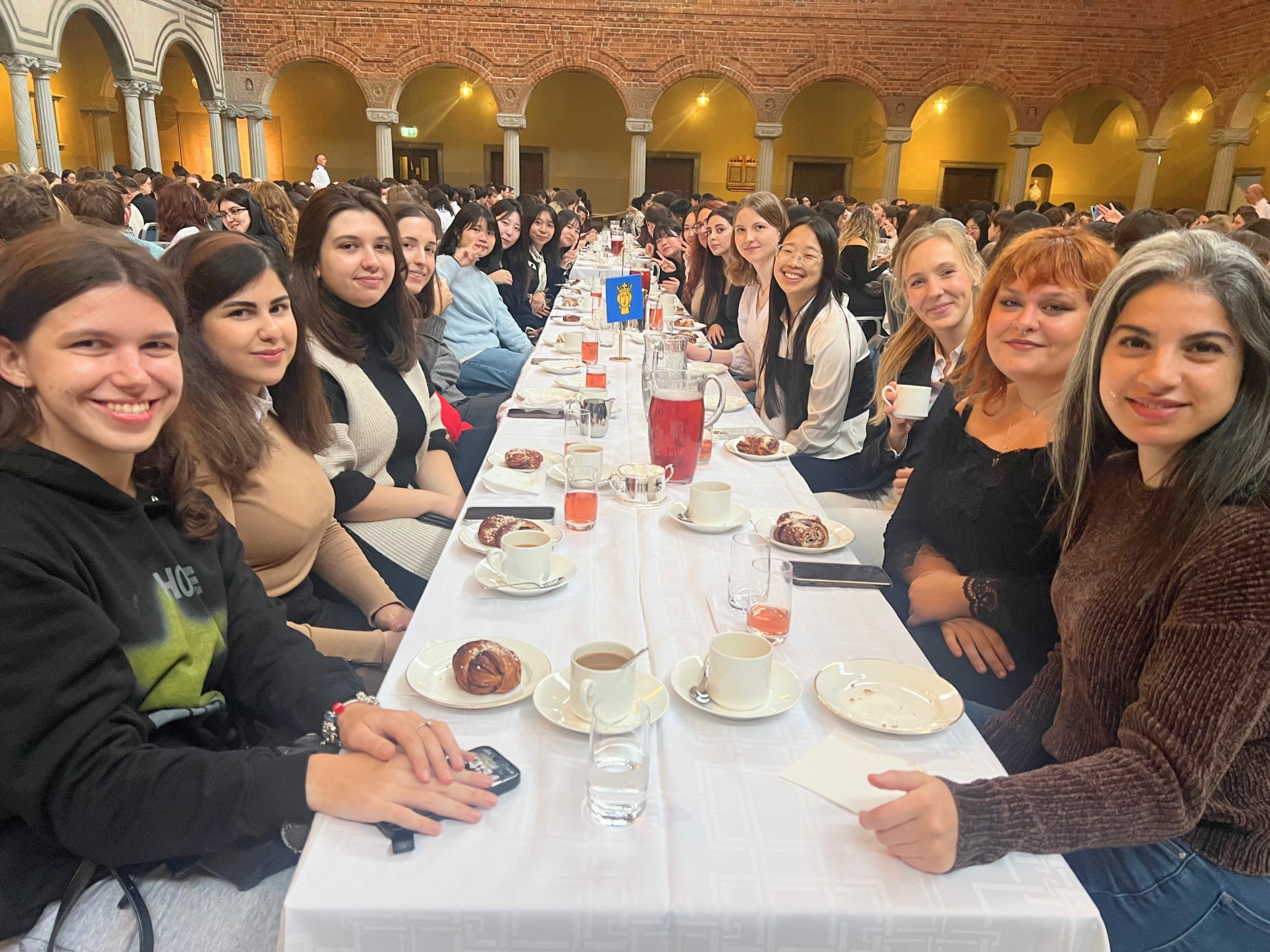 Welcome reception_City of Stockholm_STAFORUM_2 A group of international students smiling towards the camera around a long table with coffee cups, pastries and drinks in Stockholm City Hall.